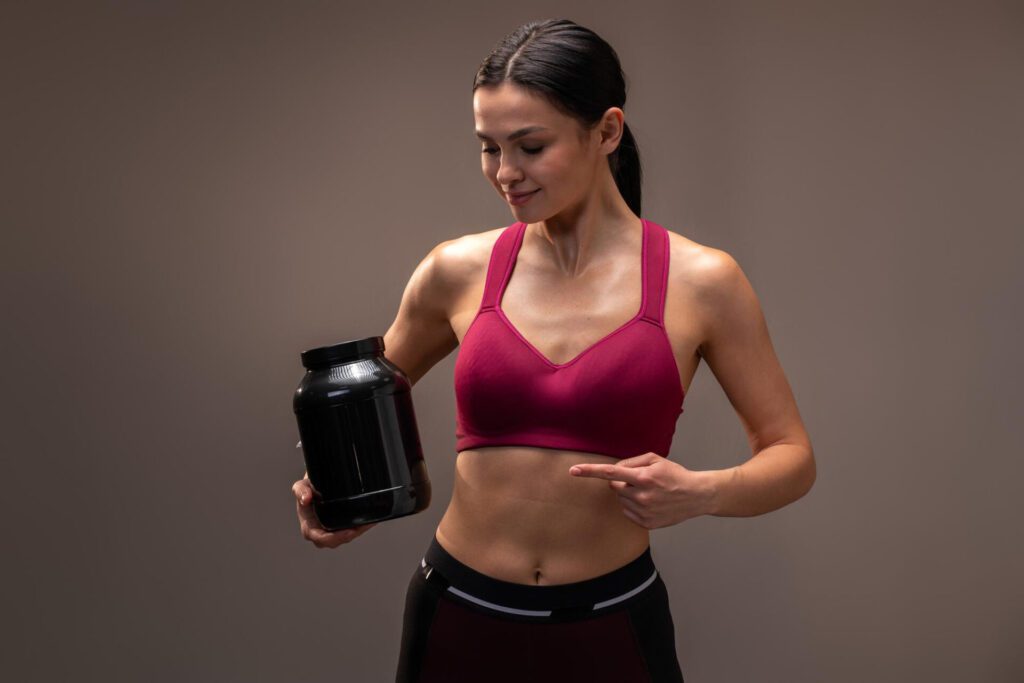 caucasian-sporty-woman-holding-shaker-with-protein-beverage-pointing-it-while-smiling-camera-proper-nutrition-concept-stock-photo