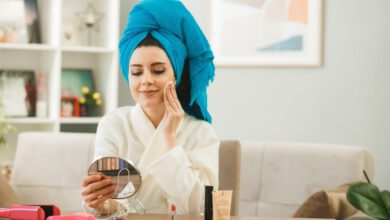 pleased young girl holding looking mirror applying tone up cream with sponge sitting table with makeup tools living room