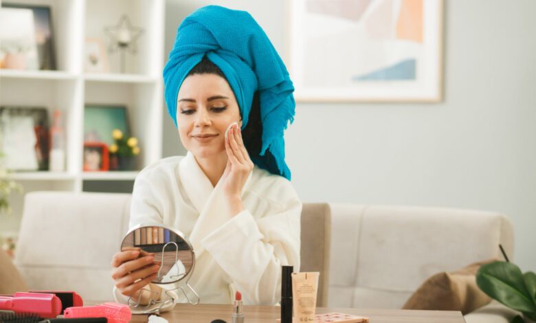 pleased young girl holding looking mirror applying tone up cream with sponge sitting table with makeup tools living room