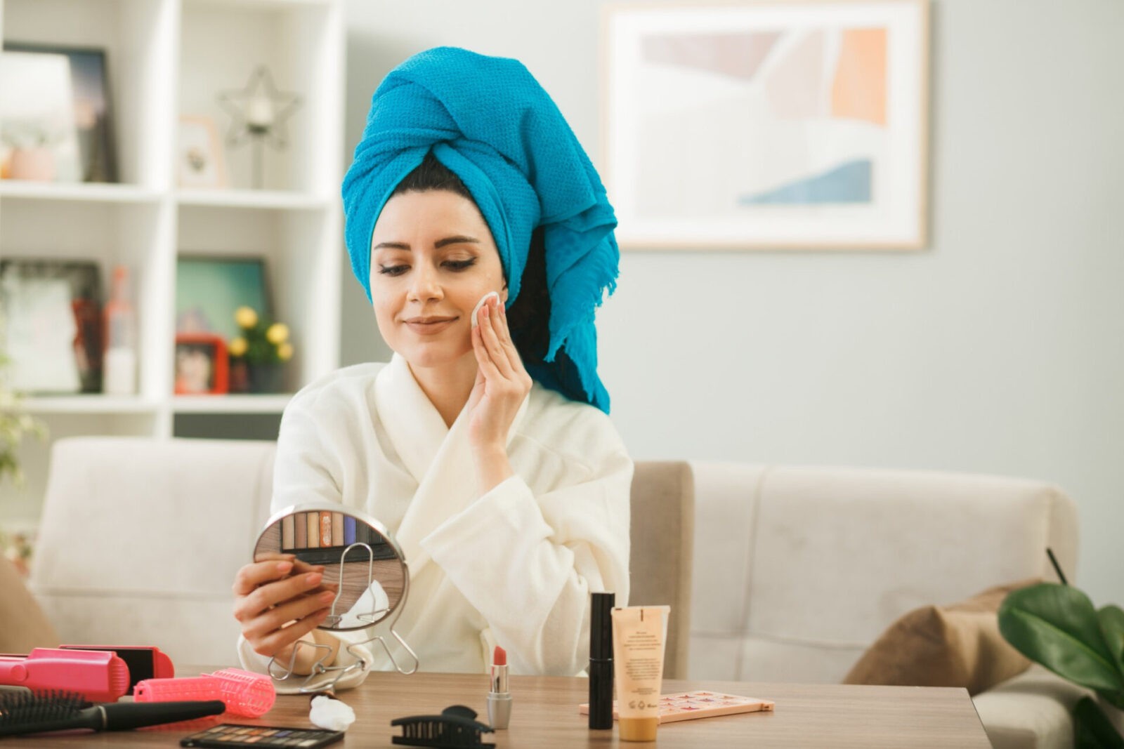 pleased young girl holding looking mirror applying tone up cream with sponge sitting table with makeup tools living room